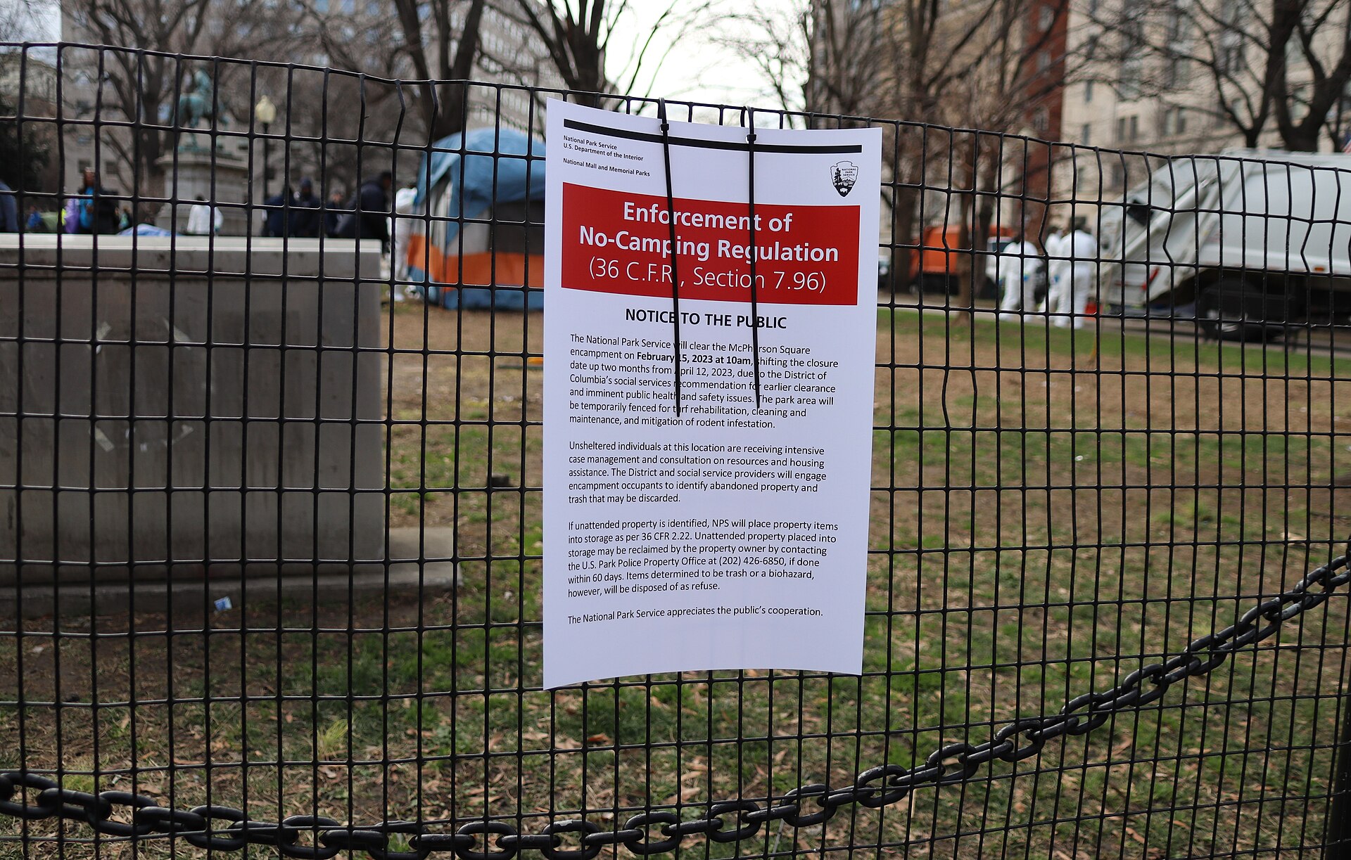A no-camping enforcement notice zip-tied to chain-link fencing over a cleared encampment
